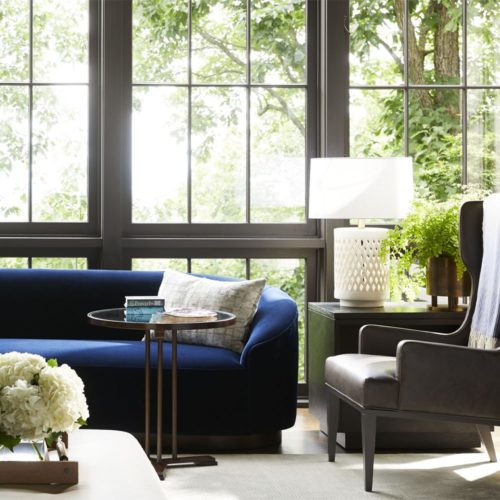 Warm and inviting open style living room with natural hues. Elegant blue velvet couch paired with leather wing chair and lovely porcelain lighting.